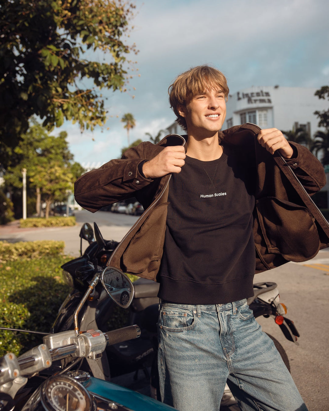 A young man smiling outdoors next to a motorcycle, wearing a black crewneck sweatshirt, a brown jacket, and light blue jeans.